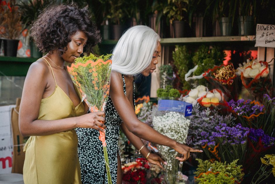 Two women shopping for flowers at a florist specializing in floral gifts on Cambridge Heath Road. The woman on the left has dark, curly hair and is wearing a yellow satin dress, holding a bouquet of o