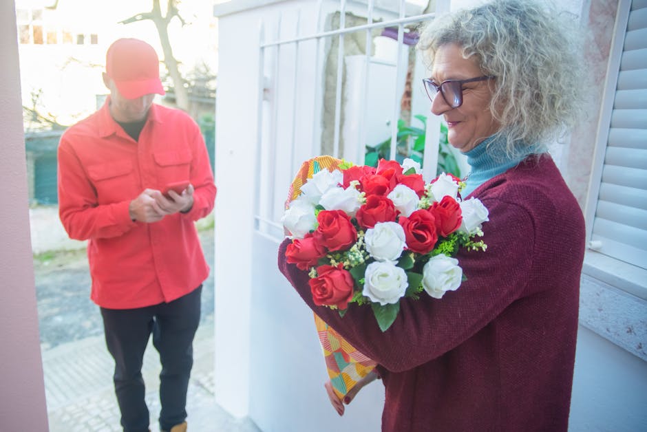 A woman with curly gray hair, glasses, and a maroon sweater holds a vibrant bouquet of fresh flowers featuring red and white roses, wrapped in colorful patterned paper. She is standing at her front do