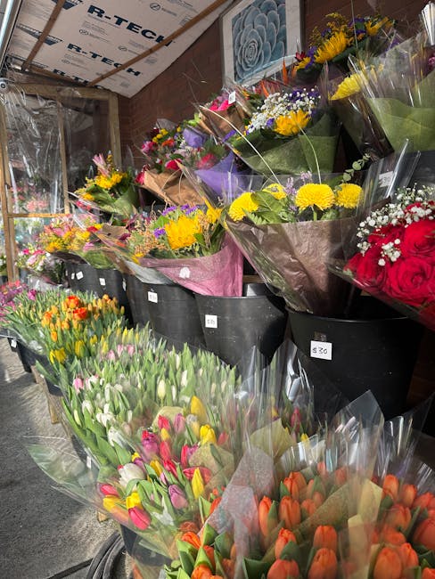 A display of various fresh flower bouquets arranged in black buckets inside a flower shop. The bouquets include bright yellow chrysanthemums, pink and white tulips, red roses, purple and white lilacs,