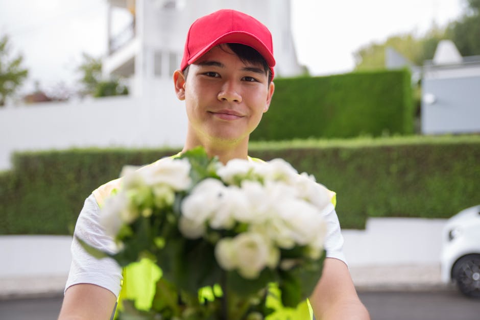 A delivery person wearing a red cap and a high-visibility yellow vest is holding a bouquet of fresh white flowers, including roses and possibly lisianthus, wrapped in clear cellophane. The flowers are
